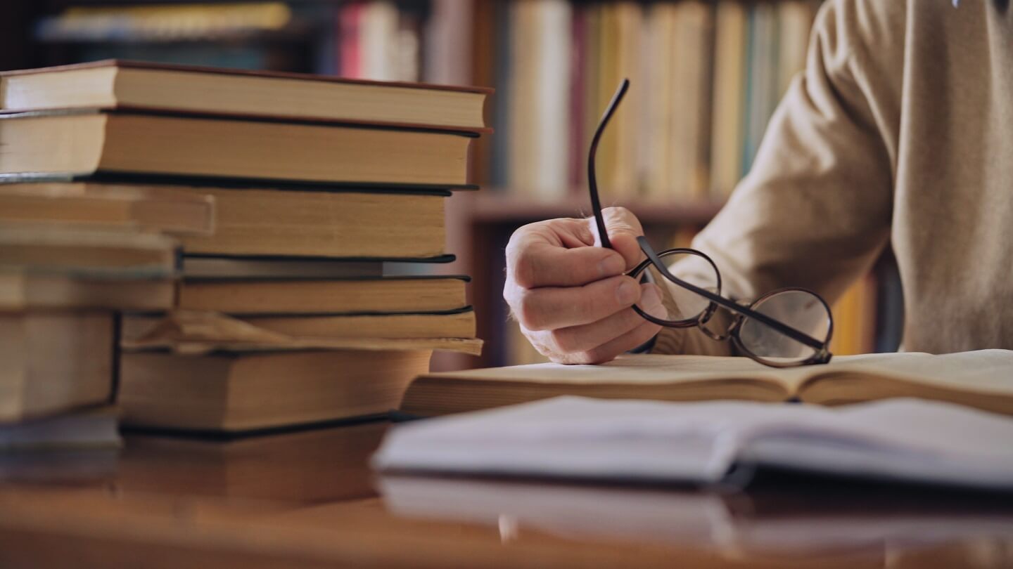 Detailaufnahme eines Mannes, der in einer Bibliothek bücher wälzt und seine Lesebrille in der Hand hält