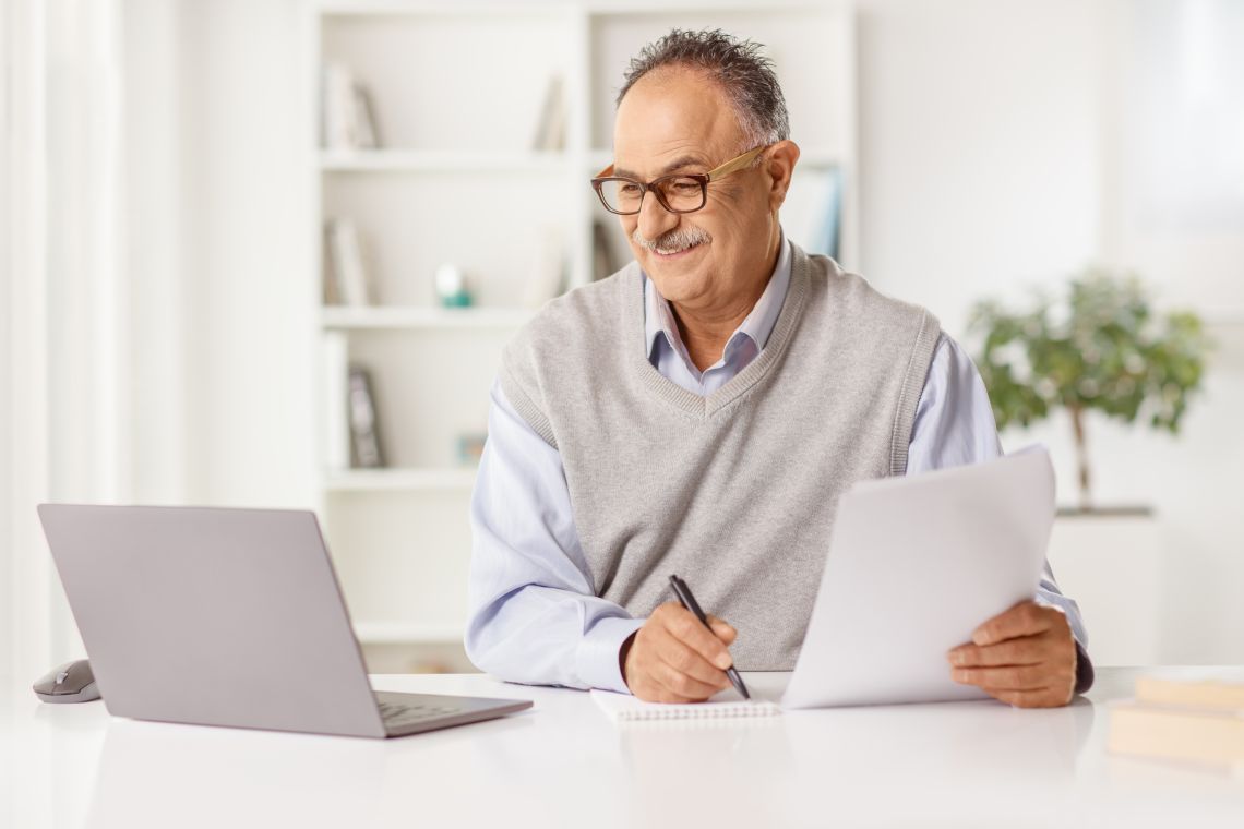 Ein Senior sitzt vor einem Laptop und macht sich Notizen