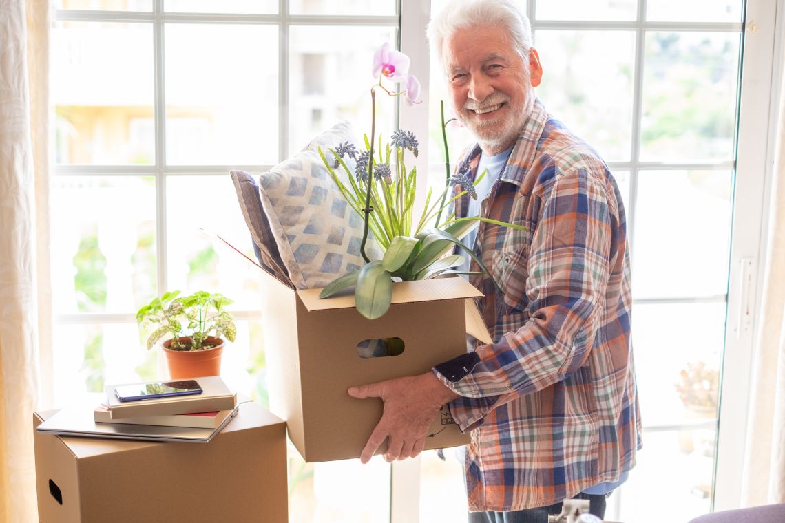 Ein lächelnder älterer Mann hält einen Umzugskarton mit Kissen und Pflanzen.