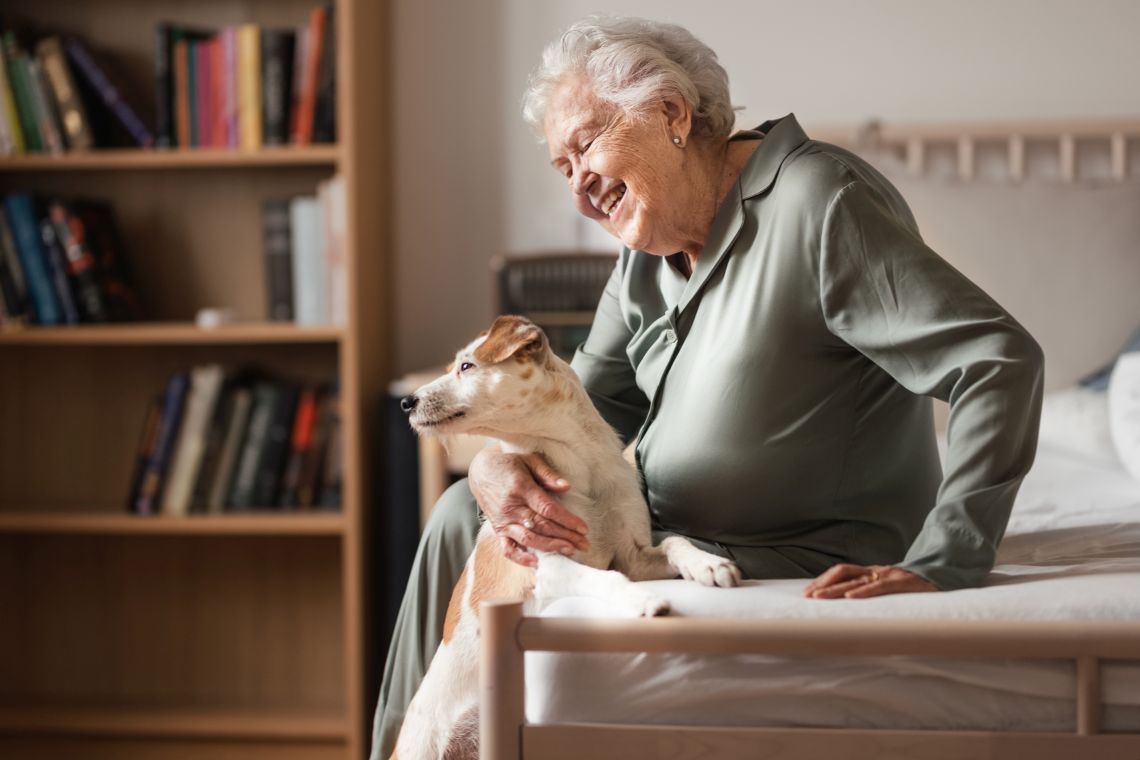 Eine Rentnerin sitzt lächelnd mit ihrem Hund auf dem Bett