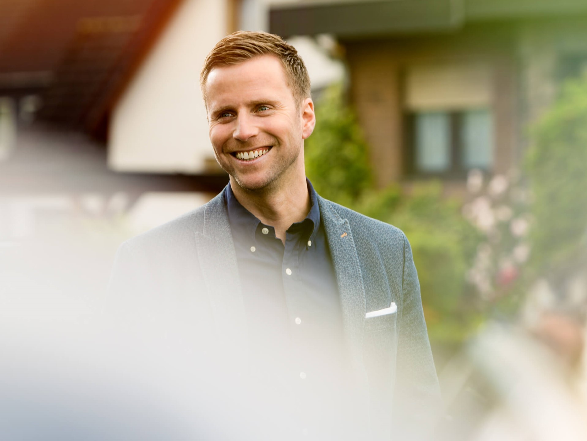 Lächelnder Mann mit kurzen Haaren der einen Blazer über einem Hemd trägt steht im Freien mit verschwommenem Grün und einem Haus im Hintergrund