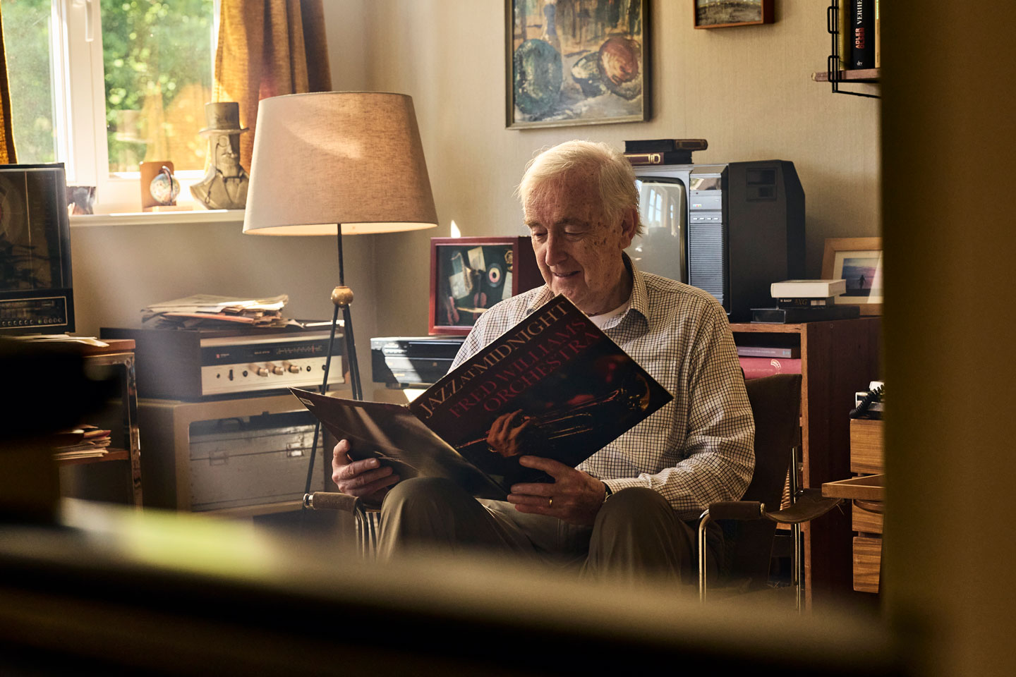 Älterer Mann sitzt in einem gemütlichen schwach beleuchteten Raum und betrachtet eine Schallplatte er ist von VintageAudiogeräten Büchern und warmem Dekor umgeben was eine nostalgische Atmosphäre schafft Sonnenlicht fällt durch ein Fenster mit halb zugezogenen Vorhängen