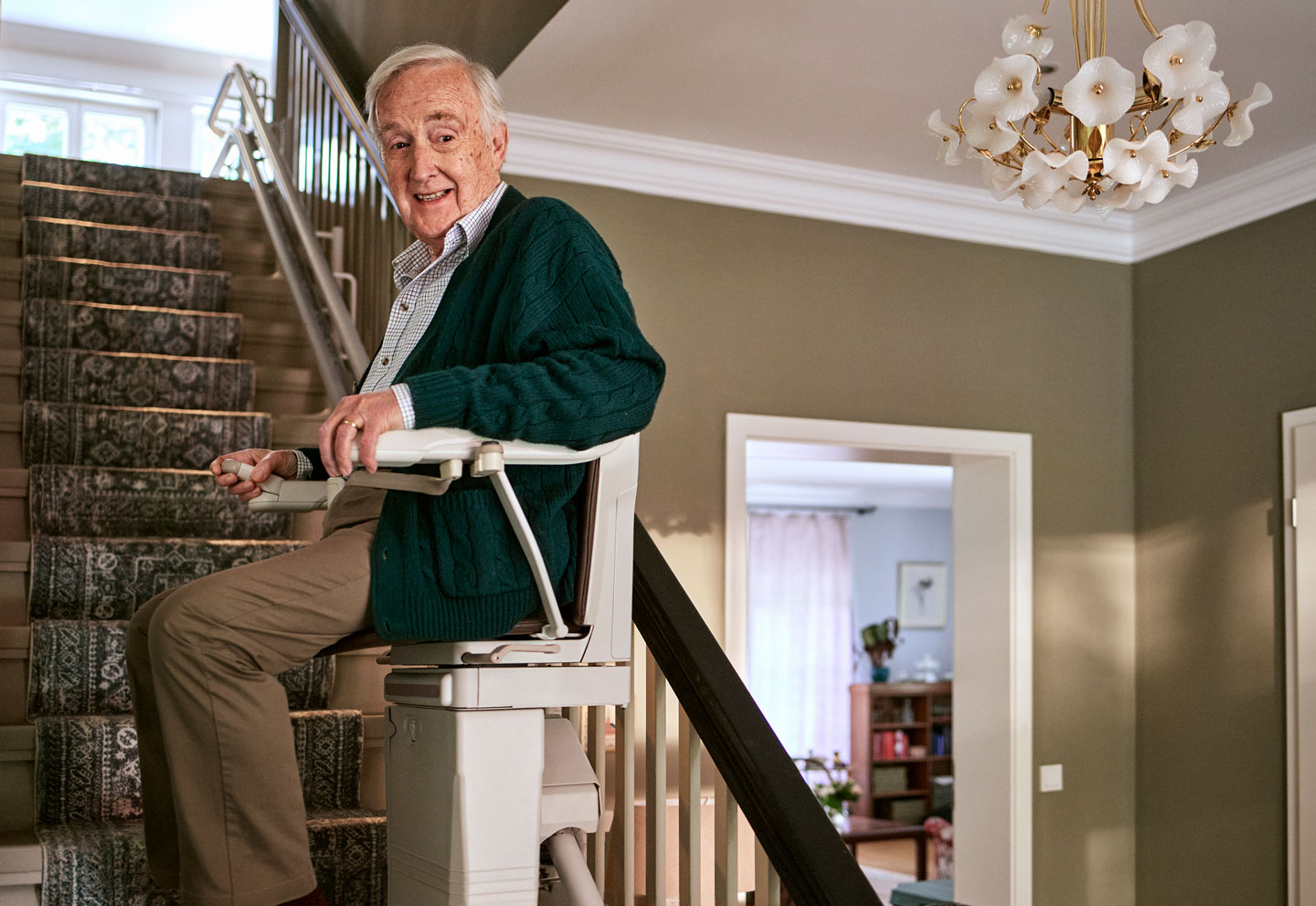 Älterer Mann mit weißem Haar der eine grüne Strickjacke und beige Hosen trägt sitzt lächelnd auf einem Sitztlift im Hintergrund ist ein gut beleuchtetes Wohnhaus mit einer Treppe einem modernen Kronleuchter und einem Blick in ein gemütliches Zimmer zu sehen