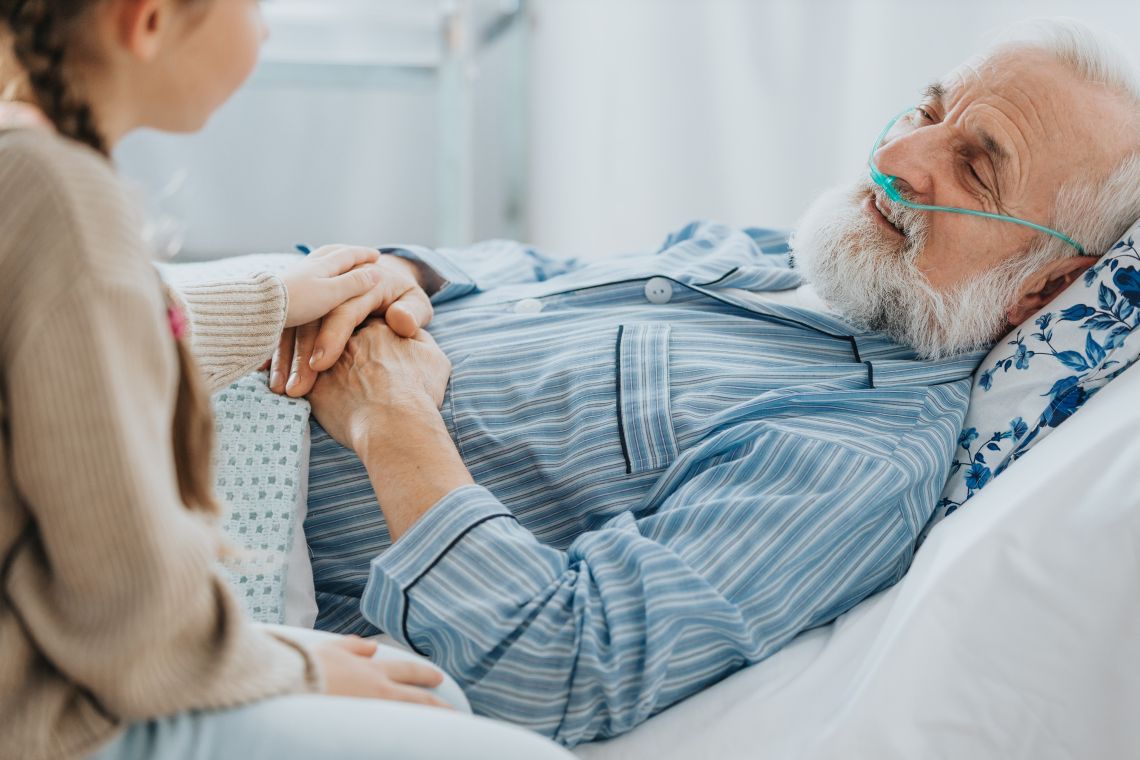 Ein alter Mann liegt im Bett, während seine Enkelin ihm über die Hand streicht.