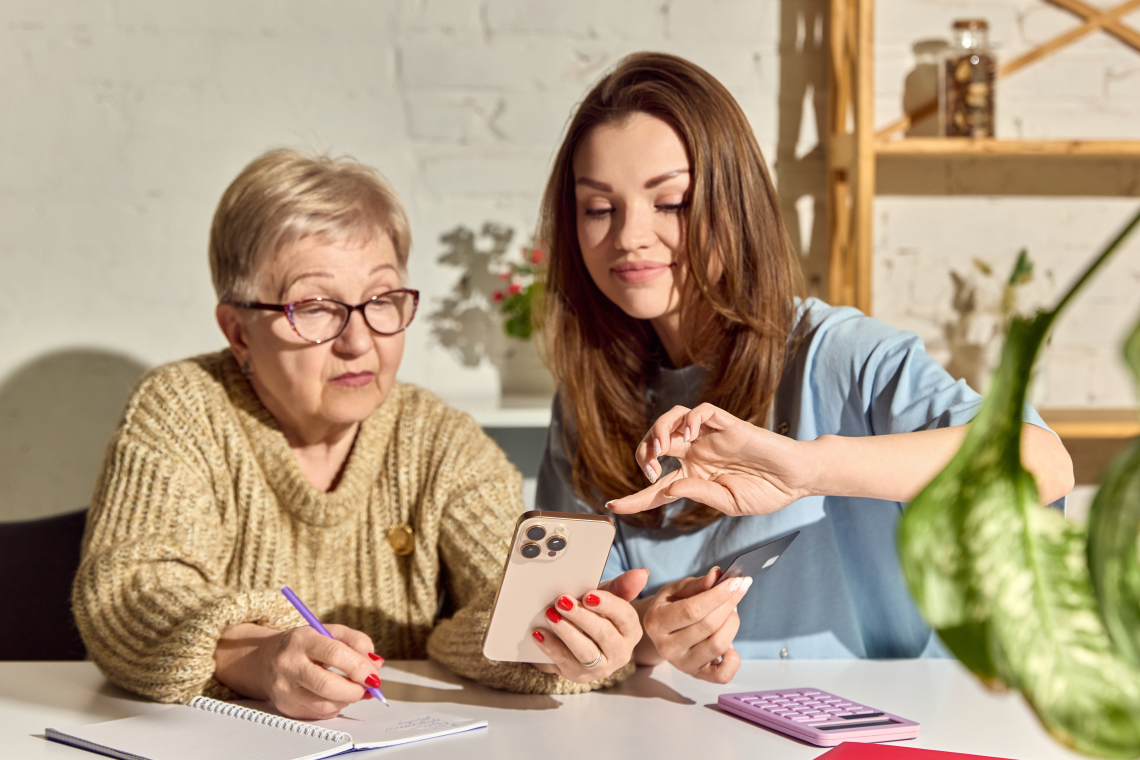 Seniorin und jüngere Frau sitzen gemeinsam am Tisch und bedienen ein Smartphone.