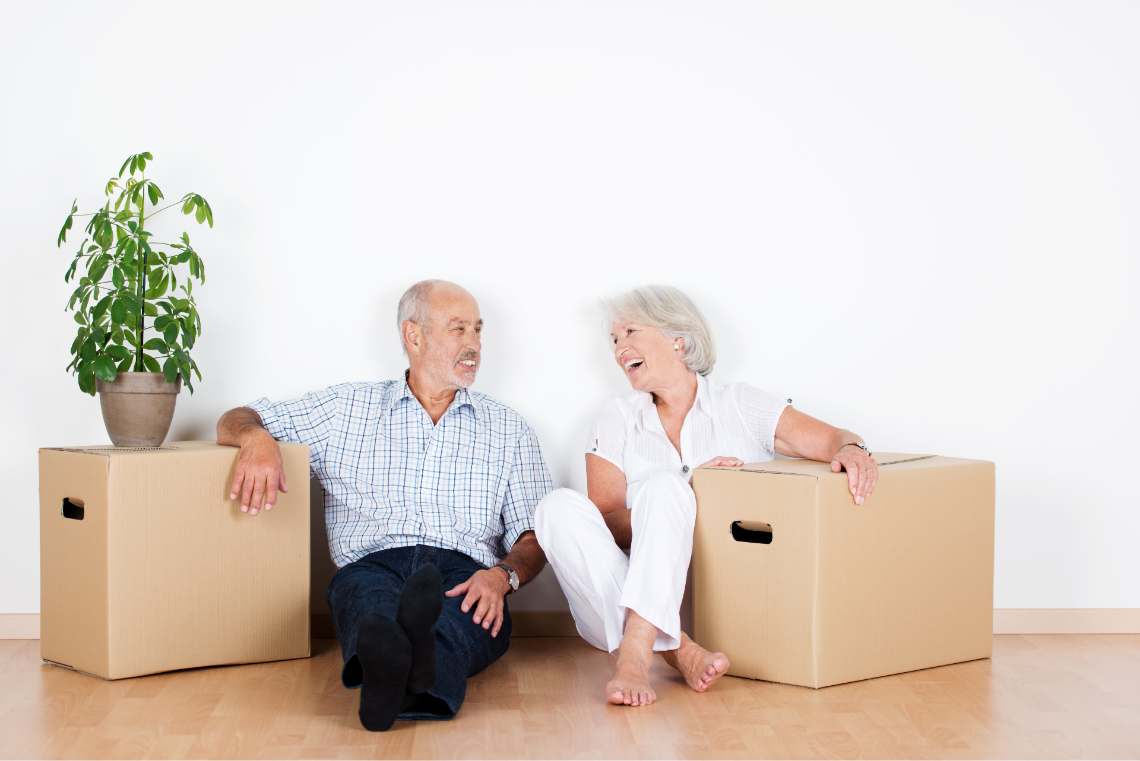 Älteres Paar sitzt mit zwei Kartons neben sich vor einer weißen Wand
