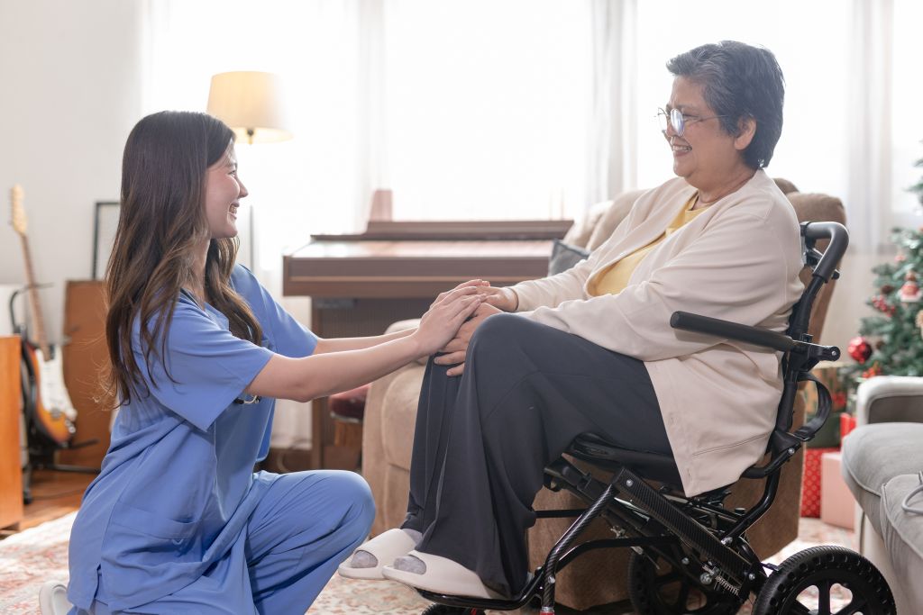 Eine junge Krankenpflegerin hält die Hände einer Seniorin im Rollstuhl und lächelt sie an
