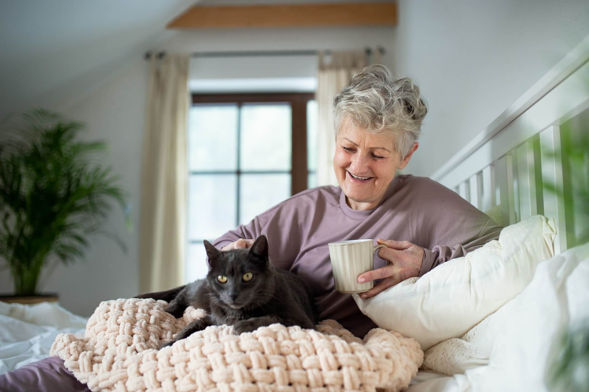 Eine ältere Dame liegt mit ihrer Katze im Bett und streichelt sie