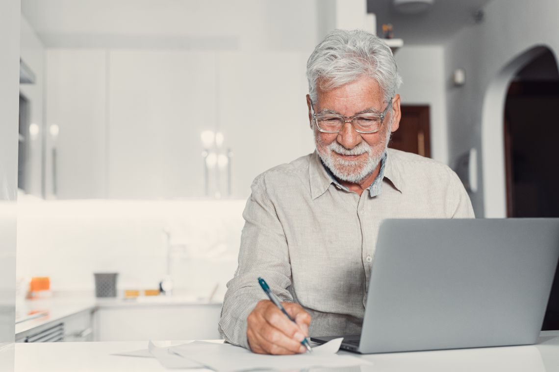 Ein Senior sitzt vor einem aufgeklappten Laptop und notiert Stellenangebote auf einem Zettel