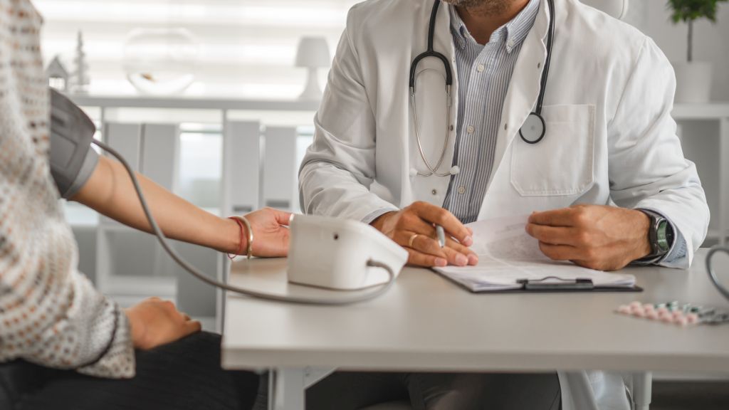Ein Arzt misst während der Sprechstunde den Blutdruck am Arm einer Patientin und notiert sich die Werte