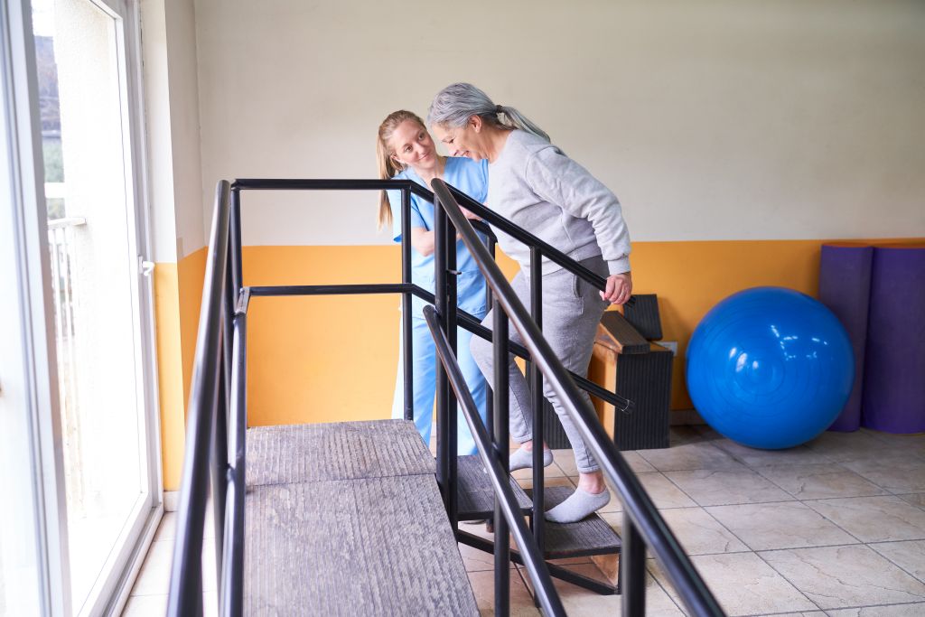 Eine Seniorin führt mithilfe einer Physiotherapeutin Übungen an einer kleinen Treppe aus, um Knieschmerzen zu vermeiden