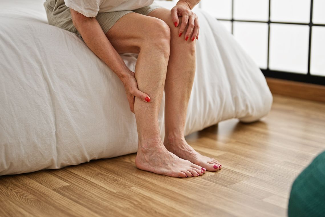 Eine ältere Person sitzt auf dem Bettrand und hält die Wade mit beiden Händen