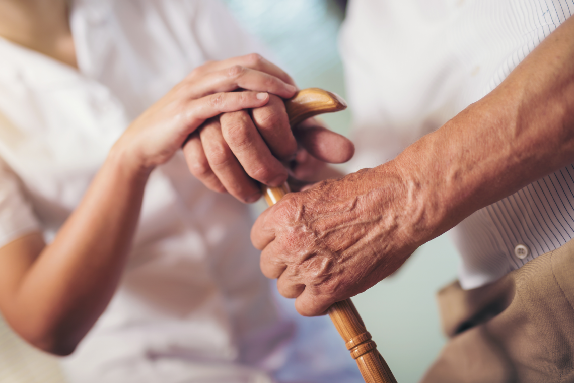 Hand einer Pflegekraft auf der Hand einer älteren Person mit Gehstock