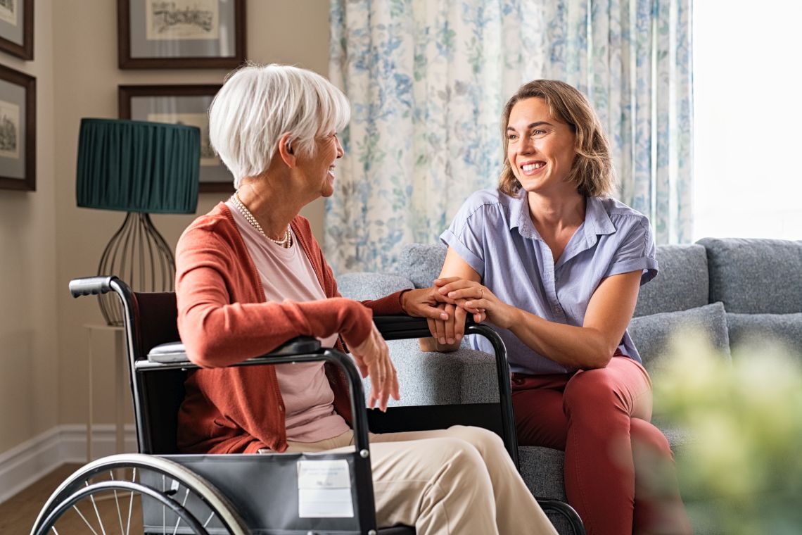 Eine Frau sitzt auf dem Sofa und hält die Hand einer Seniorin im Rollstuhl