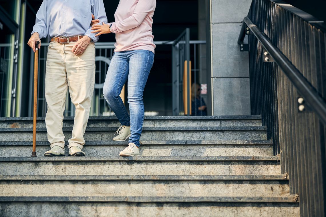 Ein älterer Mann geht mit Gehstock und einer Begleitperson die Treppe runter