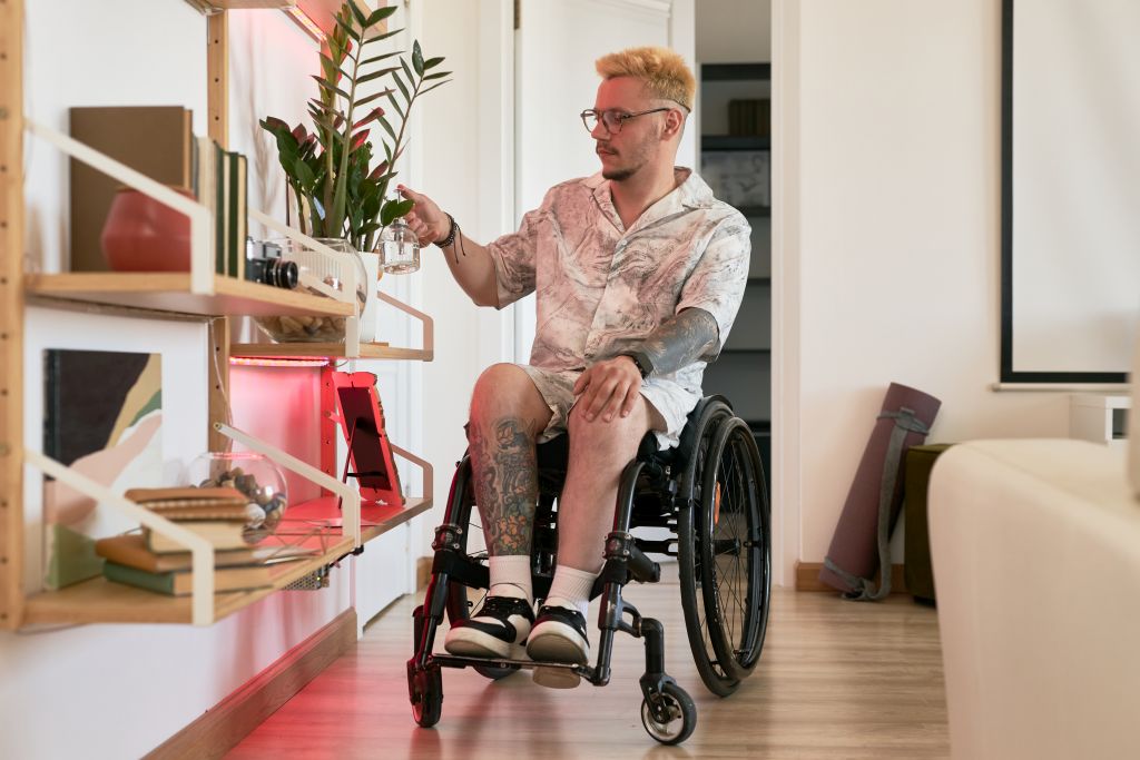 Ein junger Mann im Rollstuhl bewässert seine Pflanzen, die sich auf einem niedrigen Regal befinden