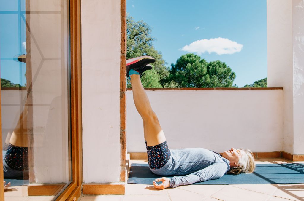 Eine Frau liegt auf einer Matte auf der Terrasse und legt die Beine an der Hauswand hoch