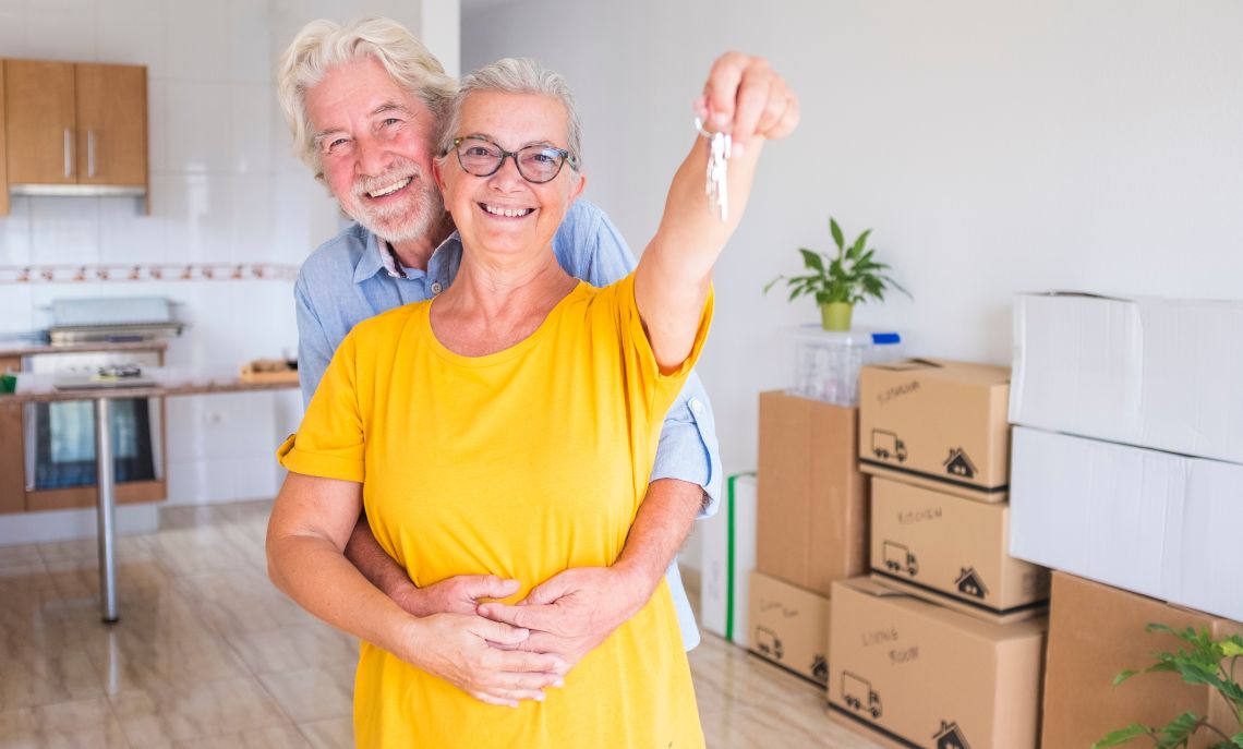 Ein Seniorenpaar steht in ihrer neuen Wohnung und umarmt sich, während die Frau den neuen Wohnungsschlüssel triumphierend hochhält.