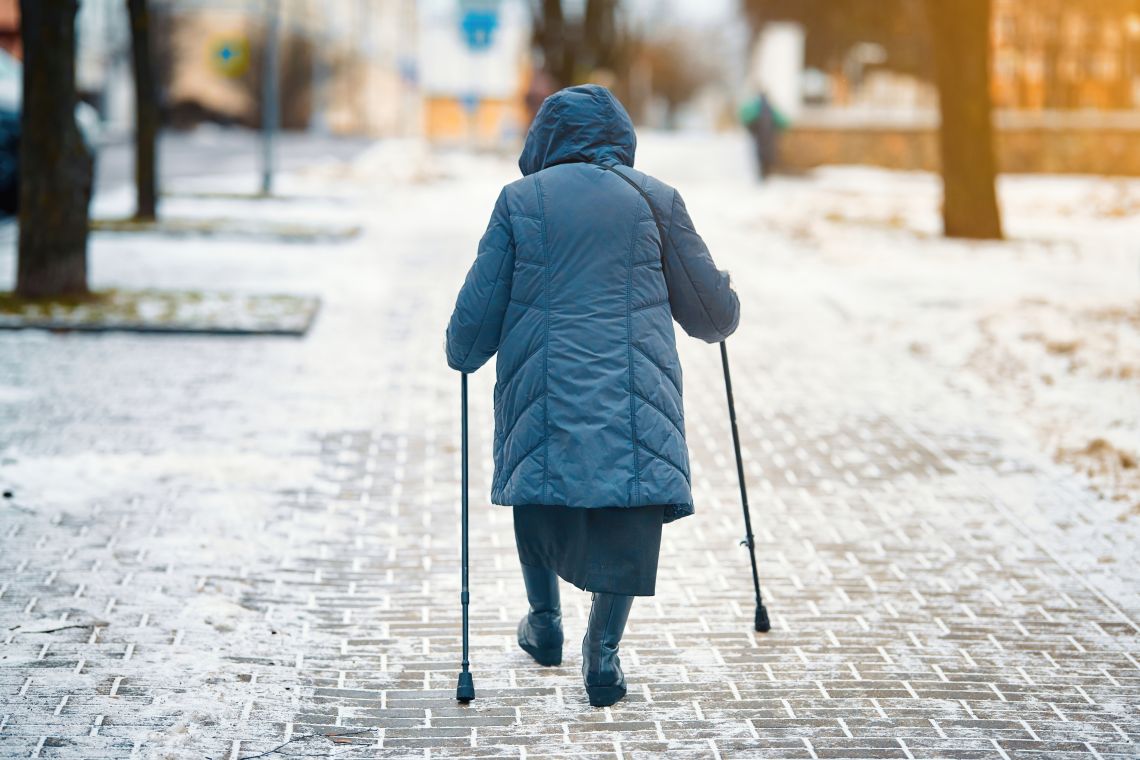 Eine Seniorin läuft im Winter mit Gehstöcken auf dem Gehweg