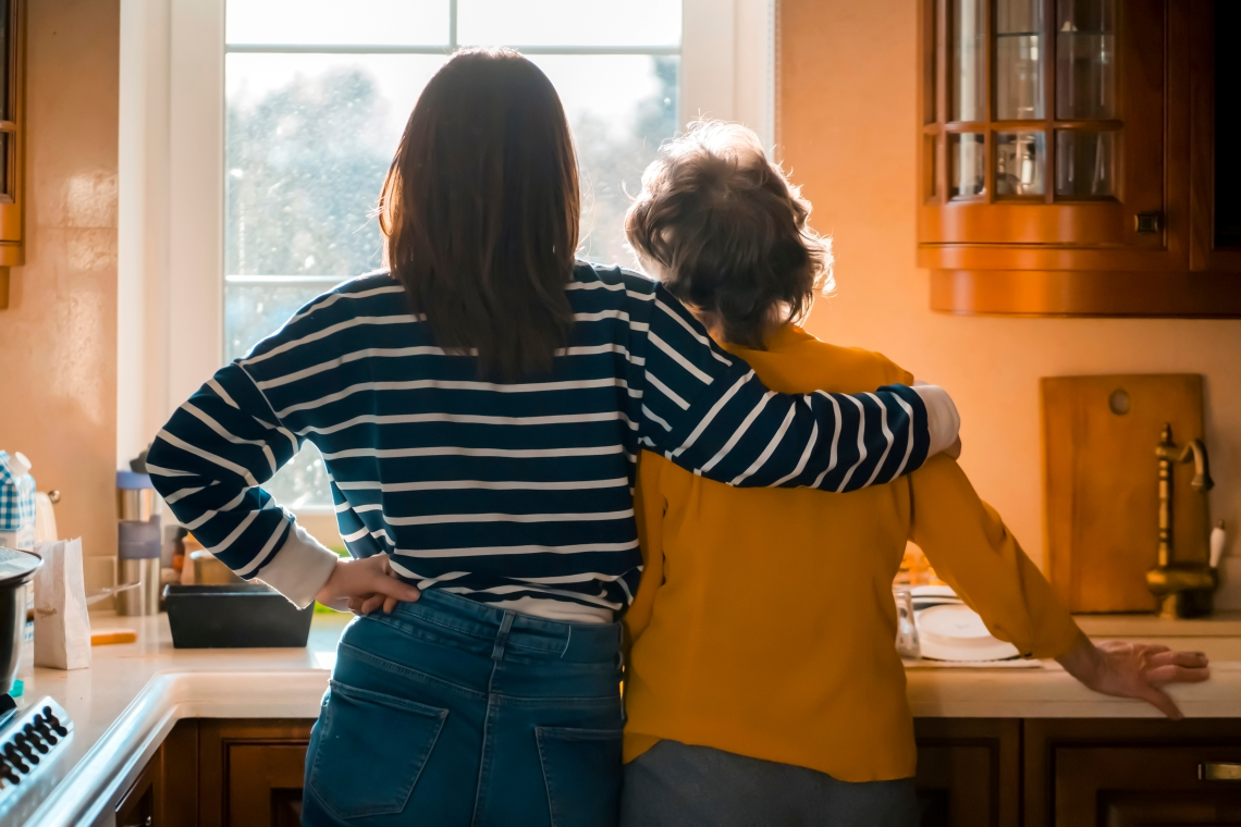 Eine erwachsene Frau steht mit ihrer älteren Mutter in der Küche. Sie halten sich liebevoll im Arm.