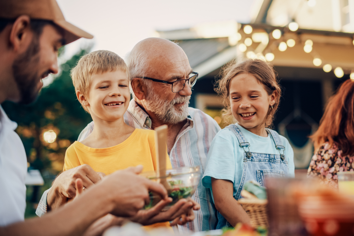 Großvater sitzt mit zwei Enkelkindern zusammen und lächelt bei einem gemeinsamen Fest im Freien.