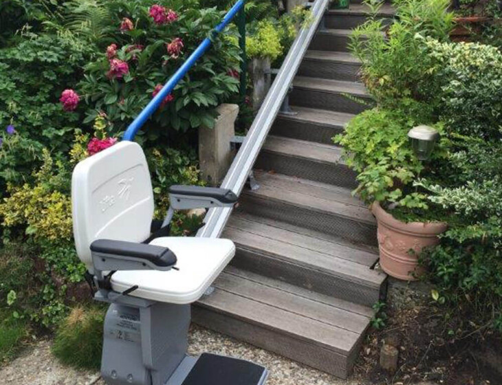 Treppenlift der an einer Außentreppe inmitten üppiger Vegetation und blühender Sträucher installiert ist und Menschen mit eingeschränkter Mobilität unterstützen soll