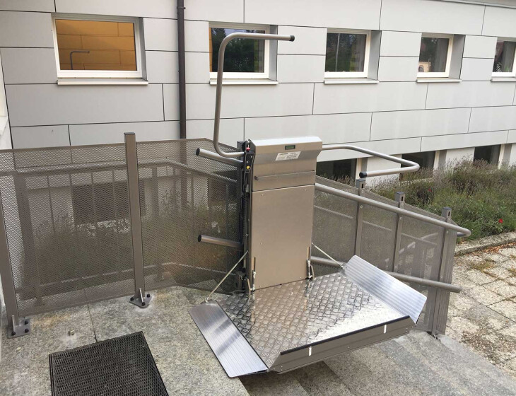 Neben einer Außentreppe ist ein Lifta Flexa installiert. Die Plattform ist hochgeklappt und mit einem Metallgeländer umgeben. Der Lift befindet sich neben einem modernen Gebäude mit grauer Fassade.